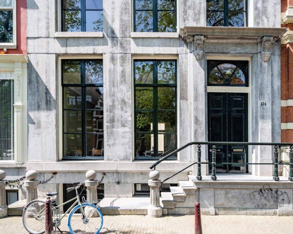 Voorgevel van een monumentaal grachtenpand aan de Herengracht 174 in Amsterdam met grote ramen, een zwarte voordeur en een fiets ervoor.