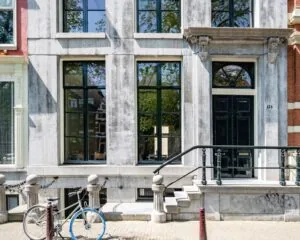 Voorgevel van een monumentaal grachtenpand aan de Herengracht 174 in Amsterdam met grote ramen, een zwarte voordeur en een fiets ervoor.