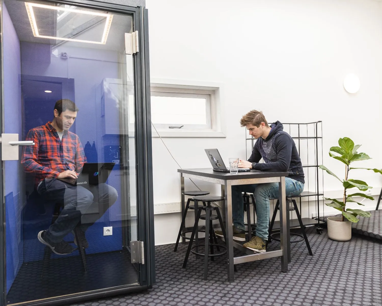 Twee mannen werken op laptops in een moderne kantoorruimte aan de Herengracht 420 in Amsterdam, één in een belcel en één aan een tafel.