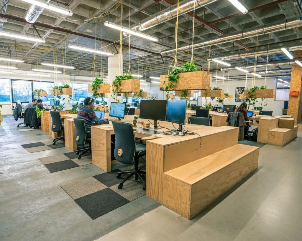 Moderne kantoorruimte aan de Johan Huizingalaan 763A in Amsterdam met houten bureaus, hangende planten en werkplekken met computers.