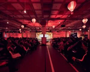 Een volle zaal met publiek kijkt naar een presentatie in een sfeervol verlichte kantoorruimte aan de Johan Huizingalaan 763A in Amsterdam.