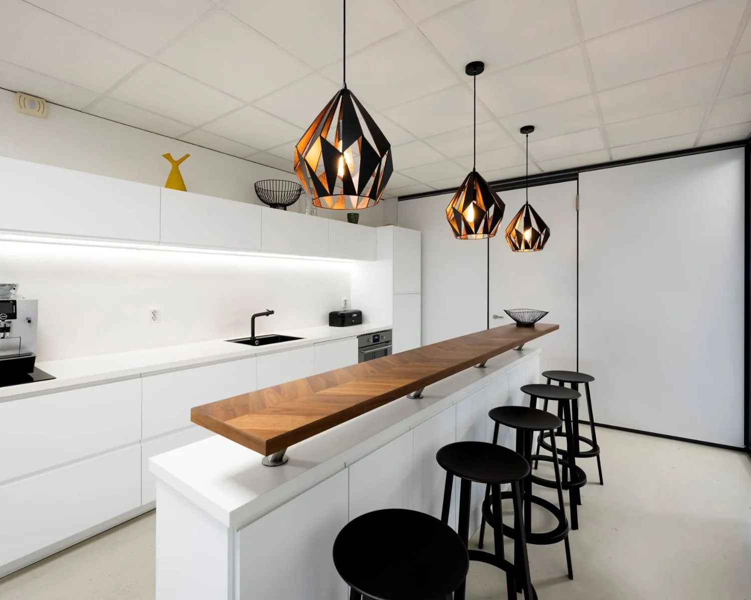 Moderne kantoorkeuken aan de Joop Geesinkweg 140-199 in Amsterdam met witte kasten, houten barblad, zwarte barkrukken en design hanglampen.