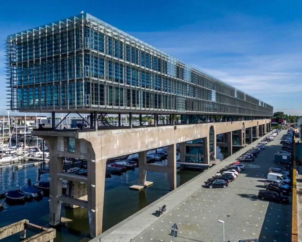 Modern kantoorgebouw Kraanspoor 12 in Amsterdam, gelegen boven een oude betonnen constructie aan het water met parkeergelegenheid eronder.
