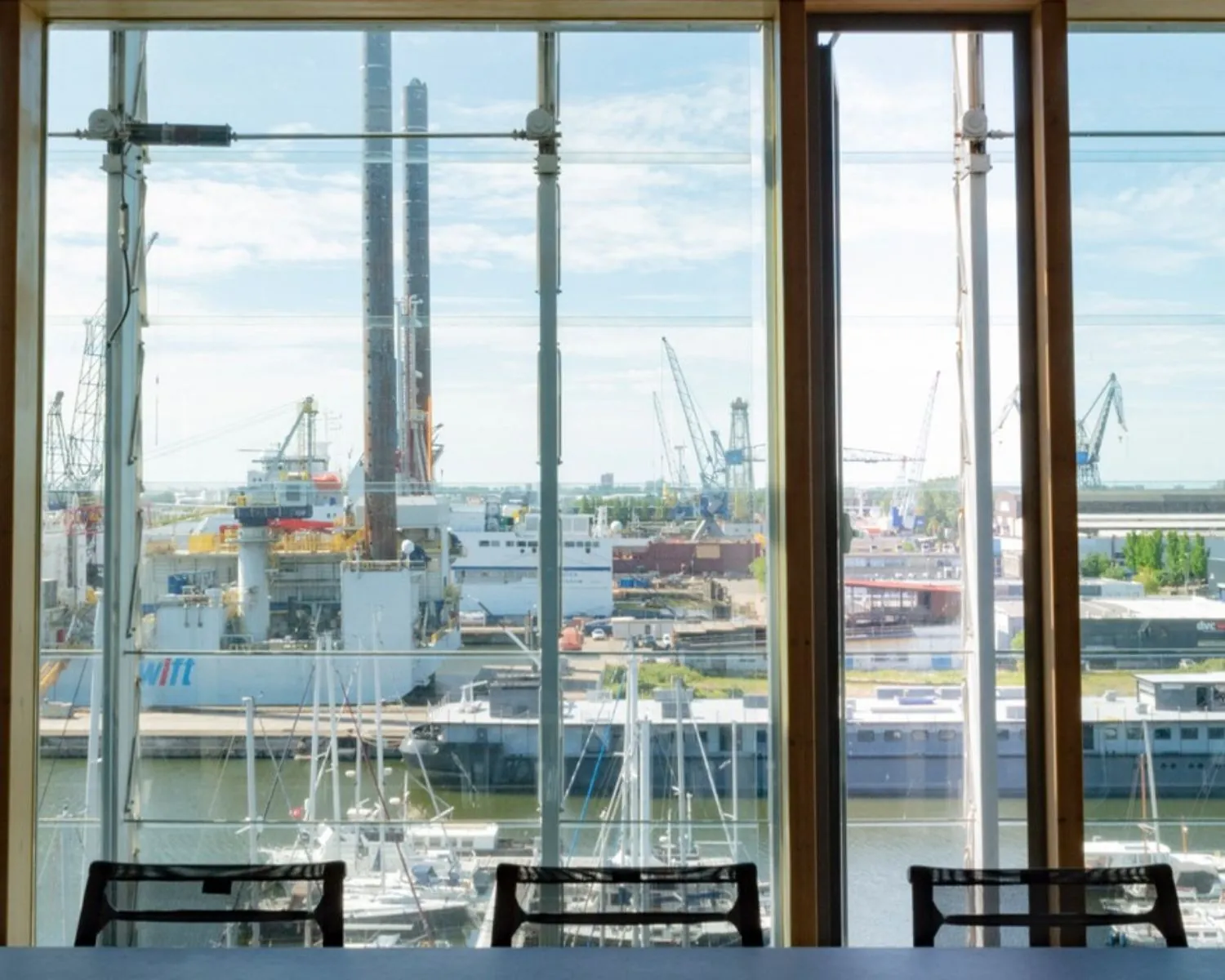 Uitzicht vanuit een kantoorruimte aan Kraanspoor 12 in Amsterdam op een haven met schepen, kranen en industriële gebouwen.