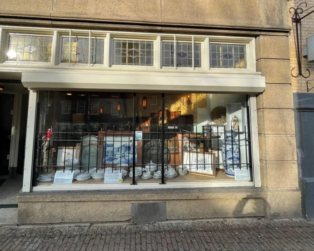 Etalage van een antiekwinkel aan de Nieuwezijds Voorburgwal 314 met serviesgoed, schilderijen en decoratieve objecten achter een tralieraam.