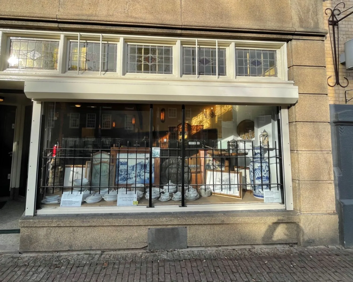 Etalage van een antiekwinkel aan de Nieuwezijds Voorburgwal 314 met serviesgoed, schilderijen en decoratieve objecten achter een tralieraam.