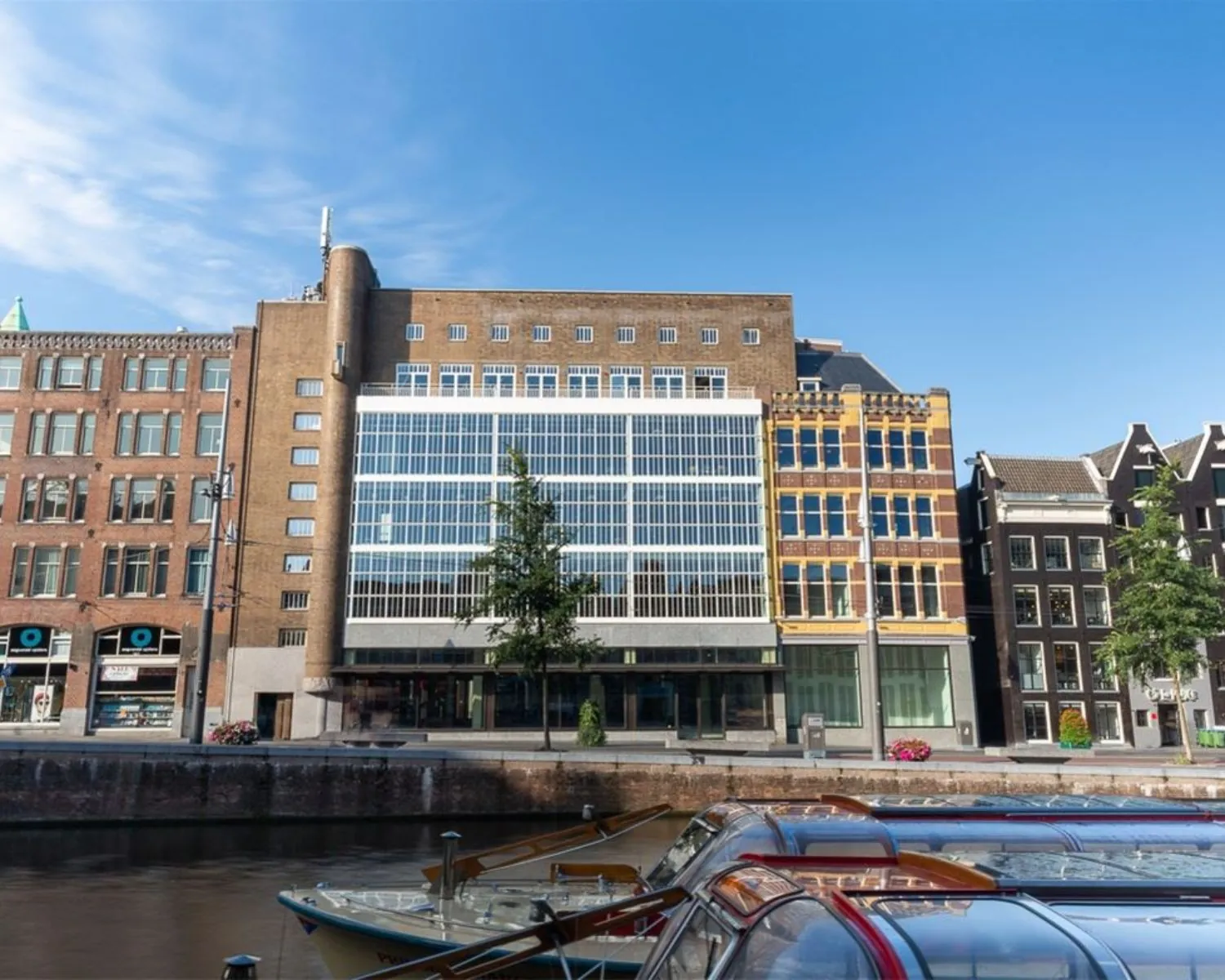 Voorgevel van een modern kantoorgebouw aan de Sint-Jorissteeg 2 in Amsterdam, gelegen aan een gracht met rondvaartboten op de voorgrond.