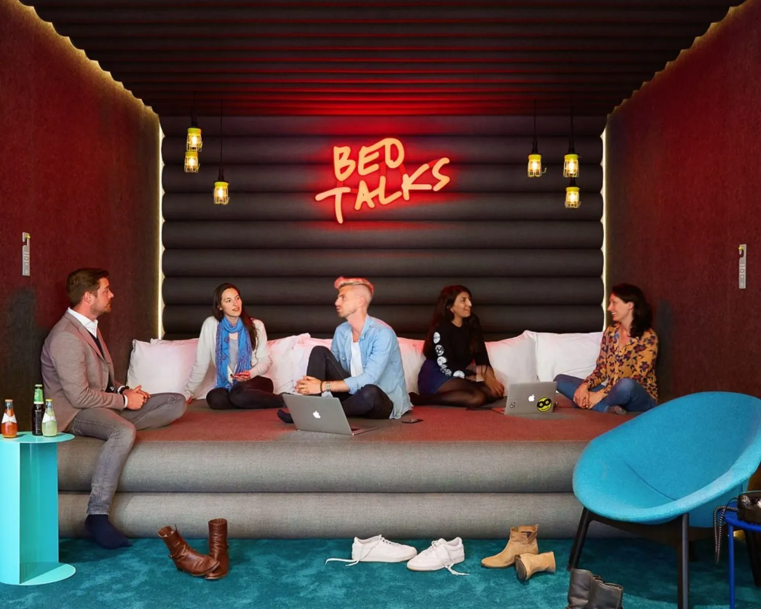 Zes mensen zitten zonder schoenen op een groot bed in een modern ingerichte kantoorruimte op Wibautstraat 131, met een neonbord "Bed Talks" op de achtergrond.