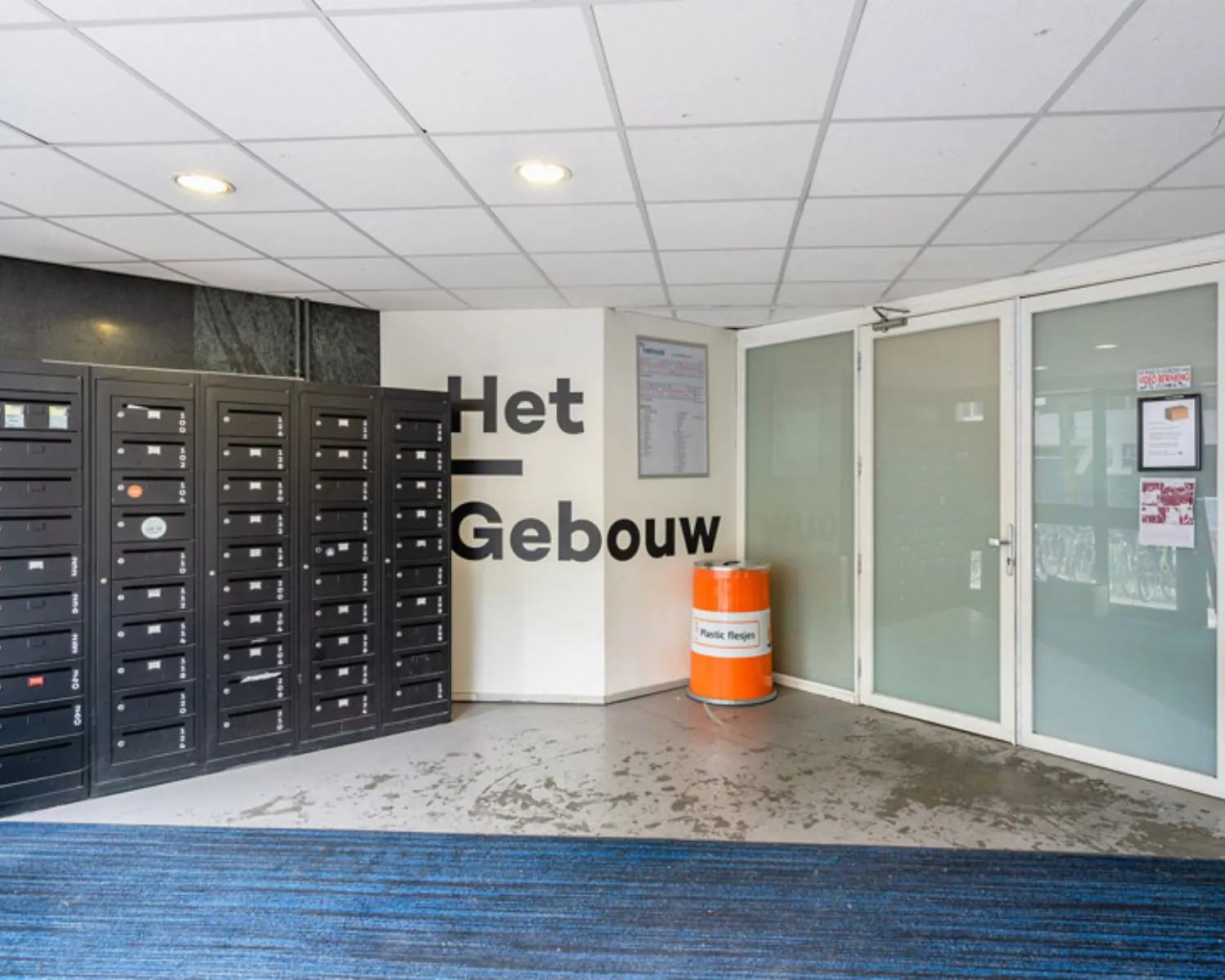 Ingangshal van Het Gebouw aan de Zomerhofstraat 71 in Rotterdam met zwarte postvakken, een oranje afvalbak en witte glazen deuren.