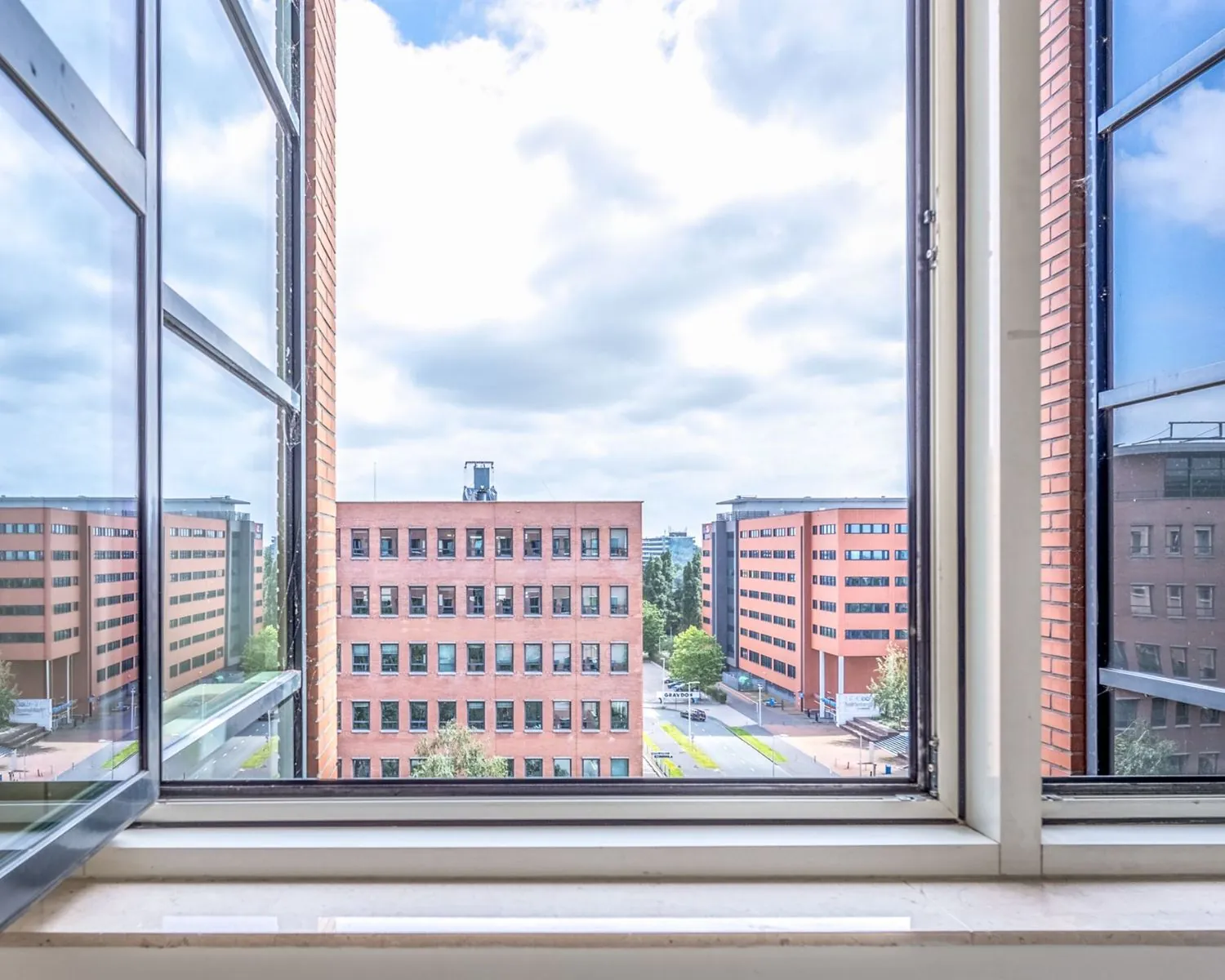Uitzicht vanuit een geopend raam op meerdere moderne kantoorgebouwen aan de Hullenbergweg in Amsterdam.