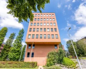 Roodbruin kantoorgebouw aan de Hullenbergweg 81 in Amsterdam, omgeven door groen en blauwe lucht.