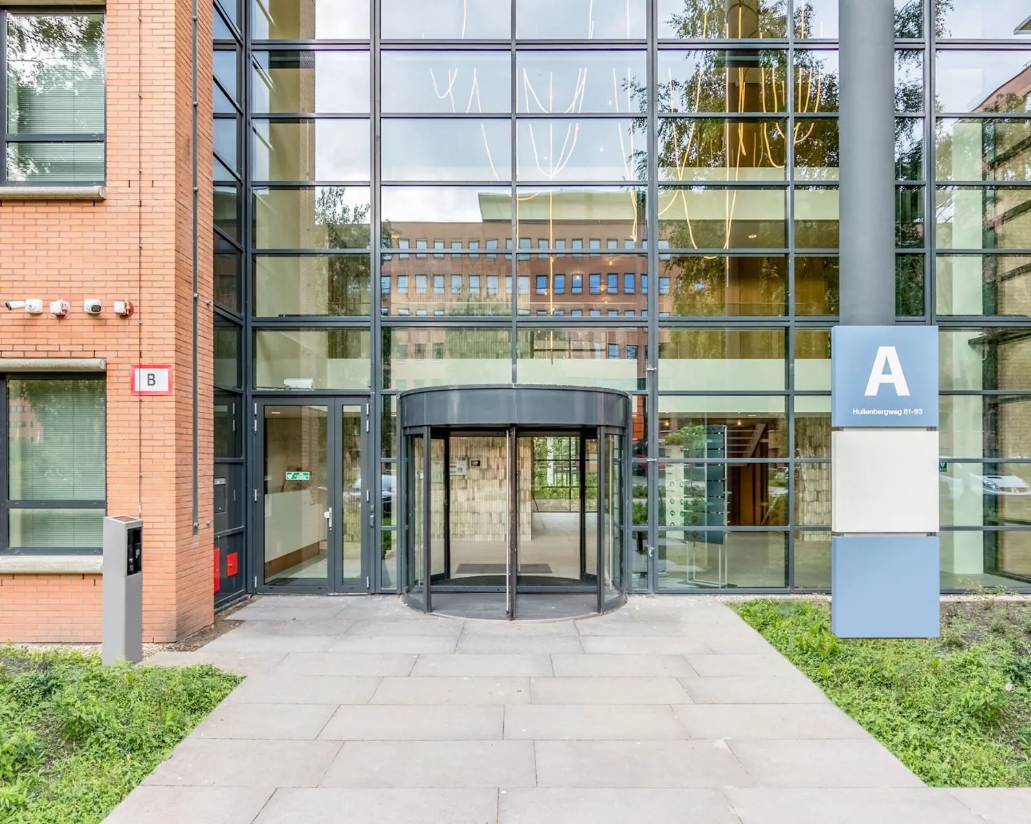 Ingang van een modern kantoorgebouw aan de Hullenbergweg 81-93 in Amsterdam met glazen gevel en draaideur.