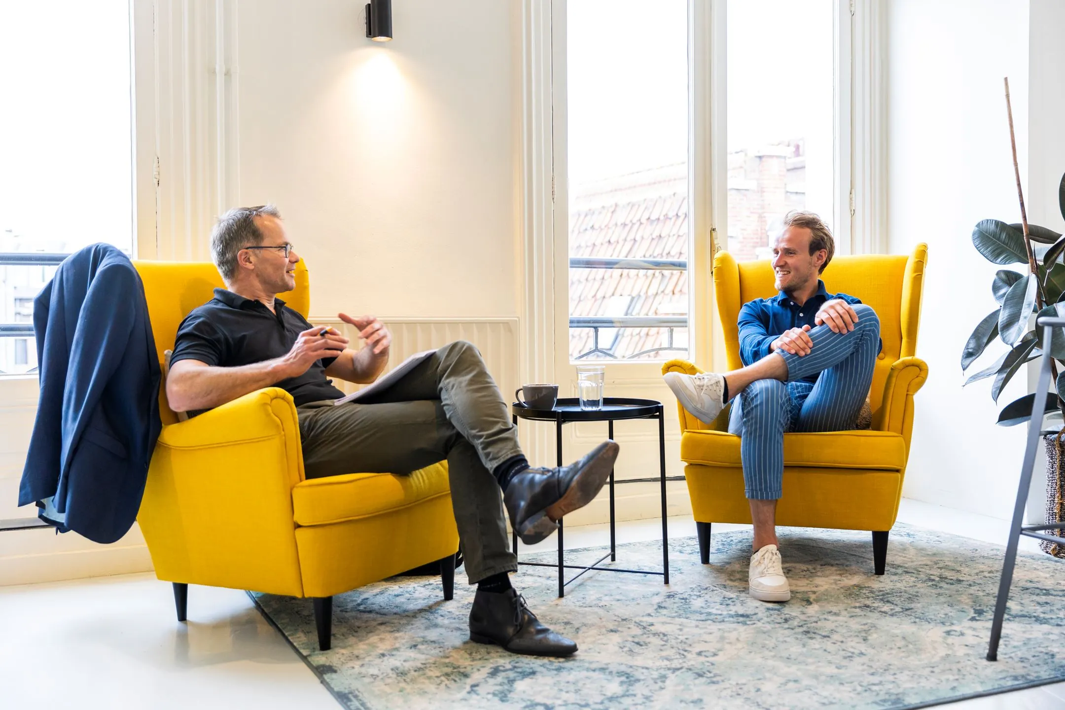 Twee mannen voeren een gesprek in een moderne kantoorruimte aan het Singel in Amsterdam, zittend in gele fauteuils bij een raam.