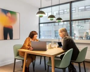 Twee vrouwen overleggen aan een houten tafel in een moderne kantoorruimte aan de Haarlemmerweg 331 in Amsterdam.