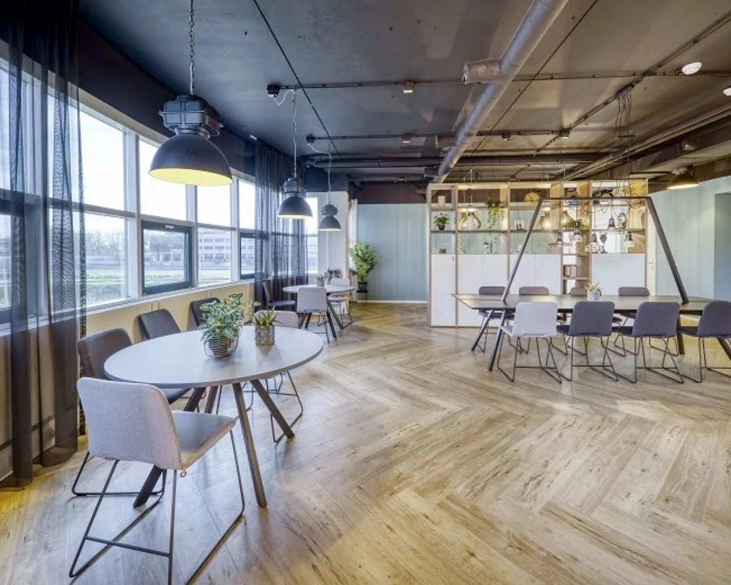Moderne kantoorruimte in Delft aan de Olof Palmeweg met ronde en rechthoekige vergadertafels, houten visgraatvloer en industriële verlichting.