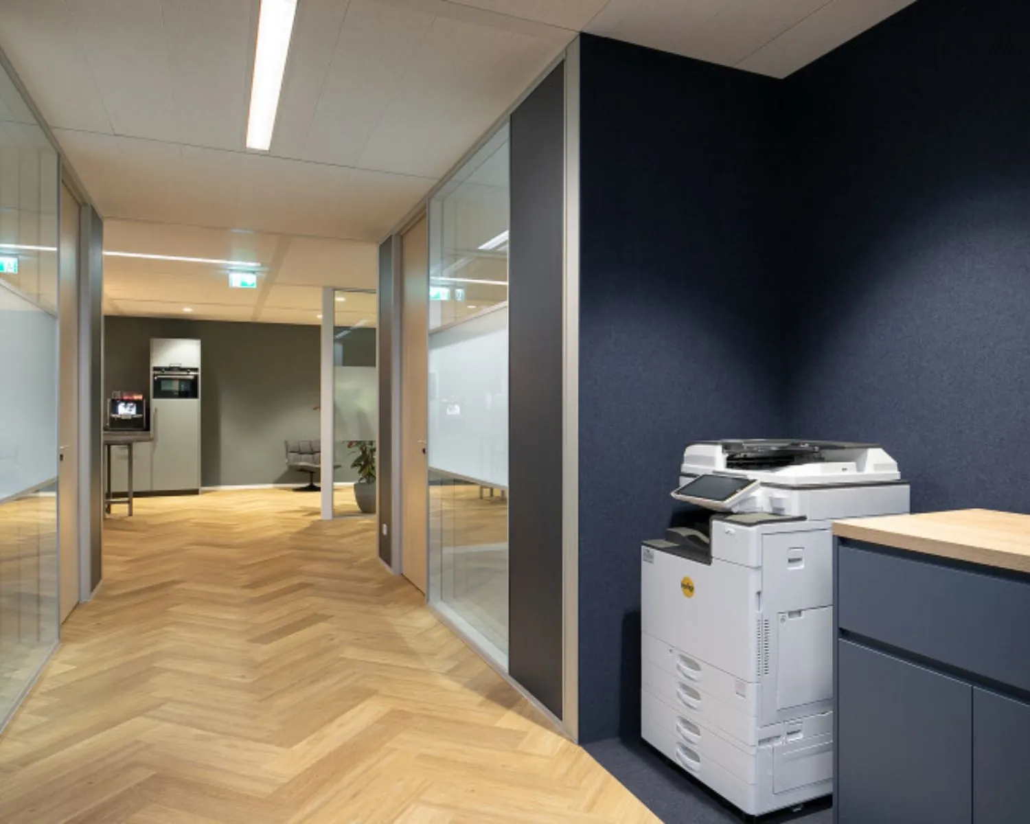 Moderne kantoorruimte in Den Haag met glazen wanden, houten visgraatvloer en een printer in de hoek.