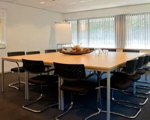 Vergaderruimte met een vierkante tafel, twaalf stoelen, flip-over en grote ramen met verticale lamellen in een kantoor aan de Herenweg 15 in Heemstede.