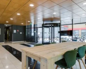 Moderne kantoorentree aan de Joop Geesinkweg 125 met houten vergadertafel, groene stoelen en een glazen draaideur.