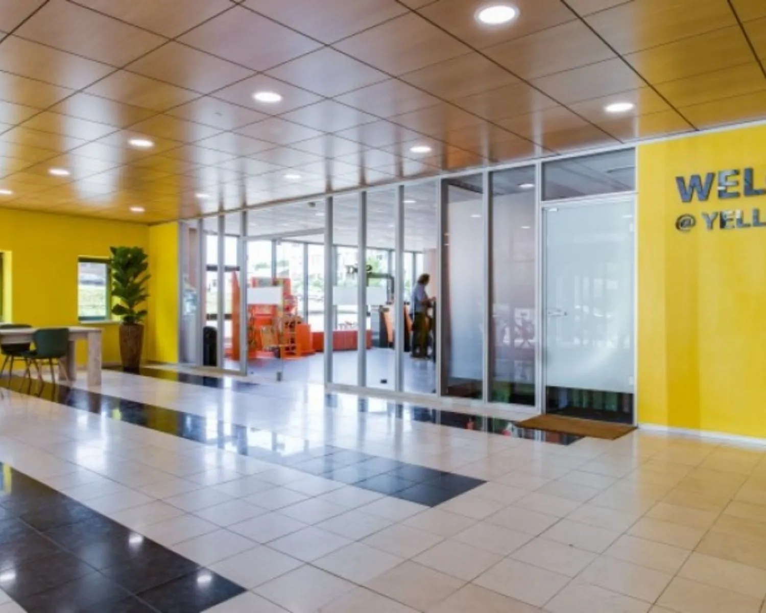 Moderne entreehal van een kantoorruimte aan de Joop Geesinkweg 125 met gele wanden, glazen deuren en een zithoek.