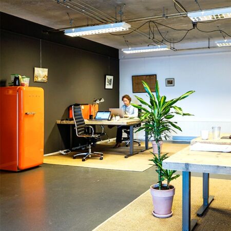 Moderne kantoorruimte met oranje koelkast, een bureau met iemand aan het werk, en diverse decoratieve elementen.