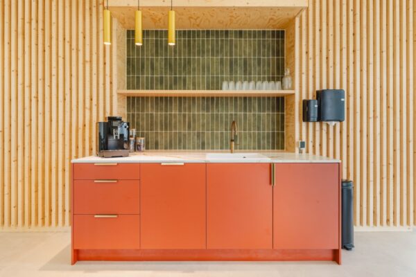 Een moderne keukenhoek met een oranje kastje, groene tegelachterwand, koffiemachine en gootsteen in een kantoorruimte aan de Basisweg 30 in Amsterdam.