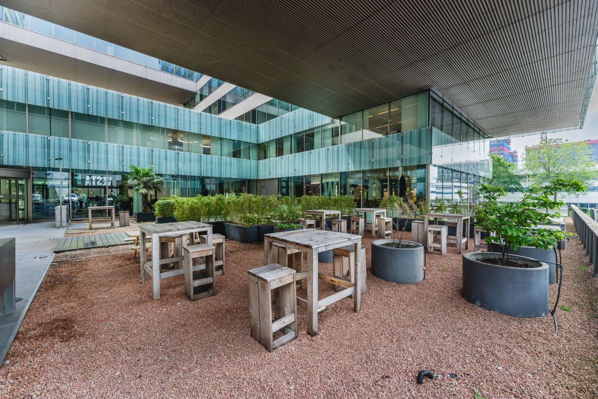 Buitenruimte met houten tafels en banken op Herikerbergweg in Amsterdam, omgeven door moderne kantoorgebouwen en planten.