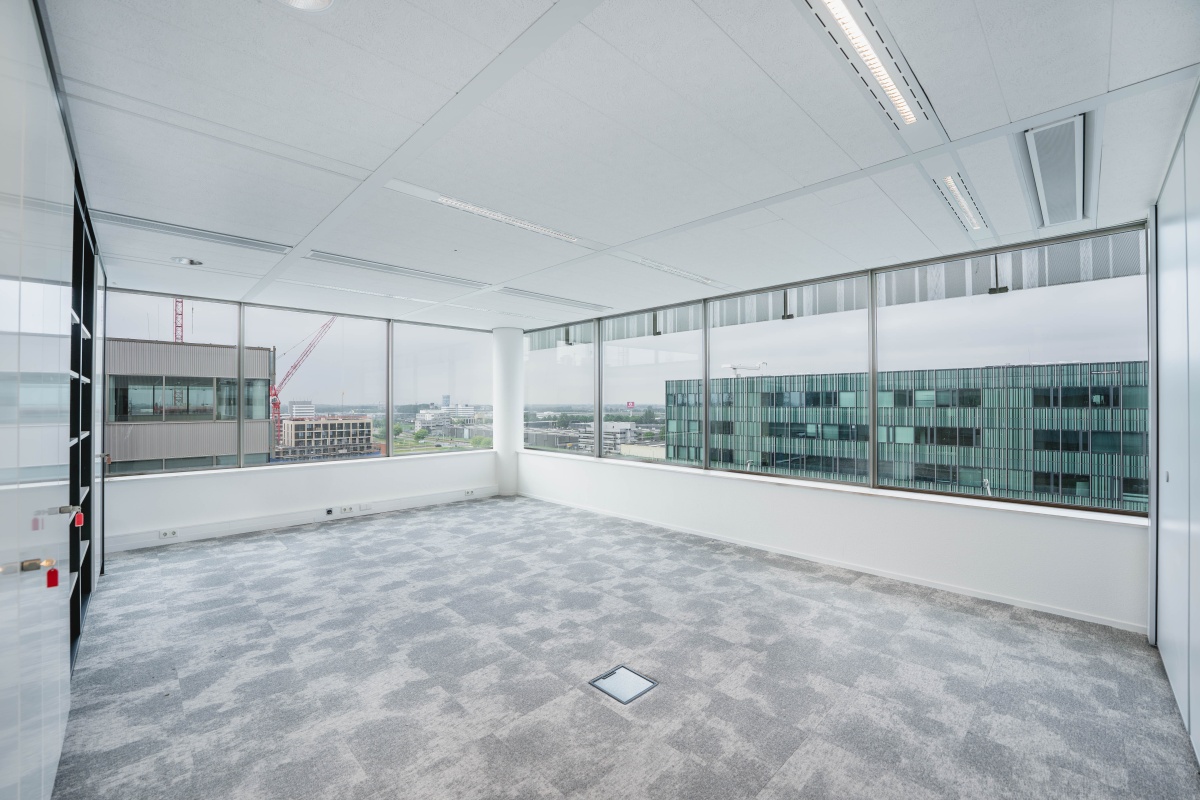 Lege kantoorruimte met grote ramen en uitzicht op gebouwen aan de Herikerbergweg in Amsterdam.