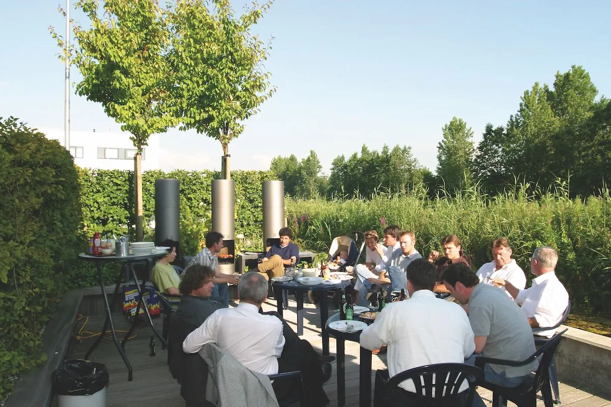 Een groep mensen zit buiten op het terras van Leeghwaterstraat 25 in Reeuwijk te eten en te overleggen in een groene, zonnige omgeving.