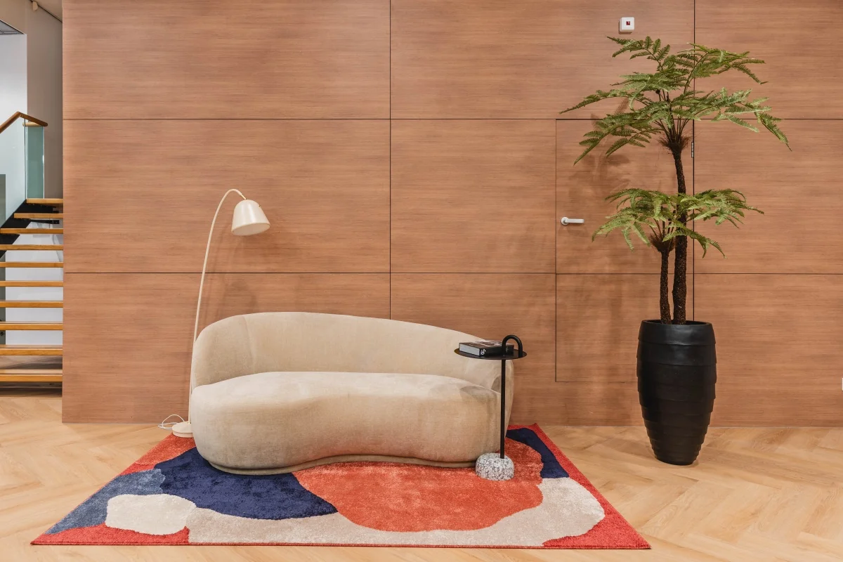 Moderne wachtruimte in kantoor Weesperstraat 107 met beige designbank, kleurrijk vloerkleed, vloerlamp en grote kamerplant.