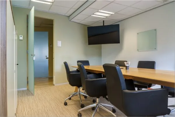 Vergaderruimte met lange houten tafel, zwarte bureaustoelen, een wandmonitor en een open deur in het kantoorpand aan Bredewater 26 in Zoetermeer.