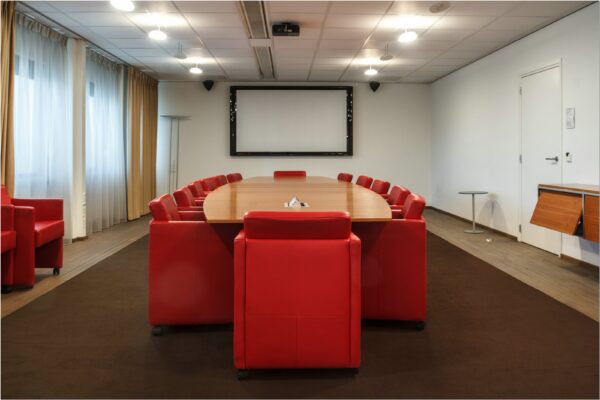 Vergaderruimte met grote ovale tafel, rode stoelen en een presentatiescherm in kantoor Bredewater 26 Zoetermeer.