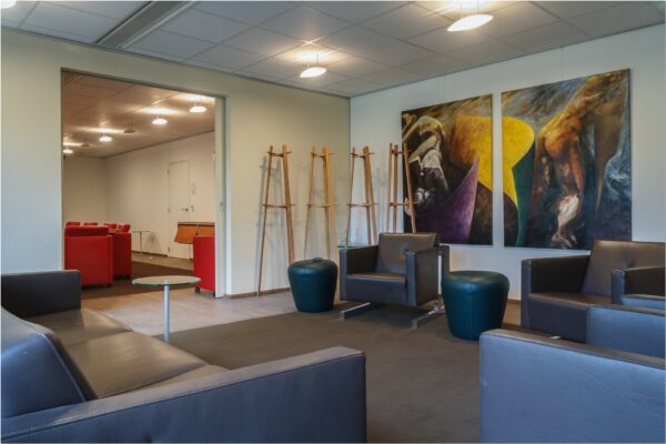 Moderne wachtruimte in kantoorruimte aan Bredewater 26 in Zoetermeer met leren stoelen, kleurrijk kunstwerk en zicht op aangrenzende ruimte met rode stoelen.
