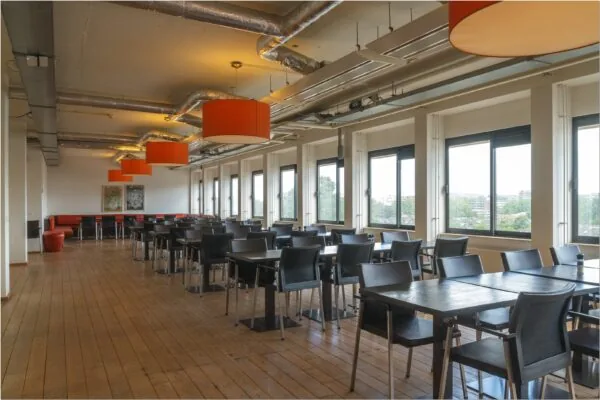 Moderne en ruime kantine in kantoorgebouw aan Bredewater 26 in Zoetermeer met grote ramen, zwarte stoelen en oranje lampenkappen.
