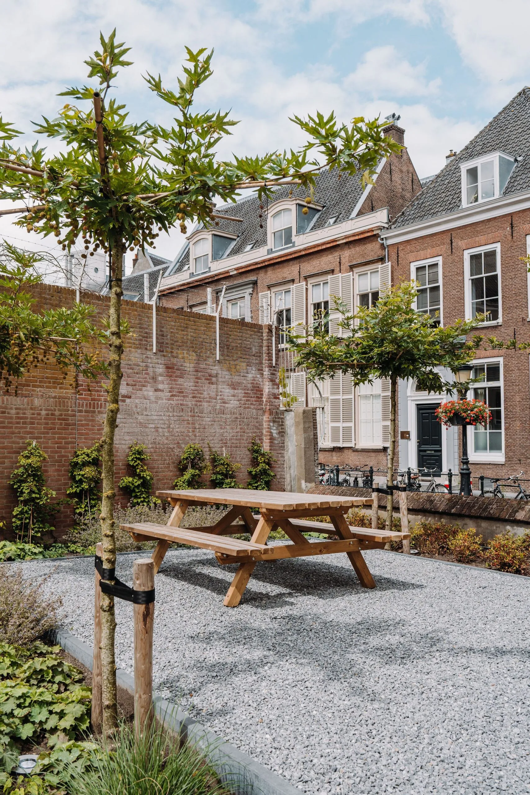 Houten picknicktafel op een binnenplaats met grind, omringd door jonge bomen en historische panden aan de Kromme Nieuwe Gracht in Utrecht.