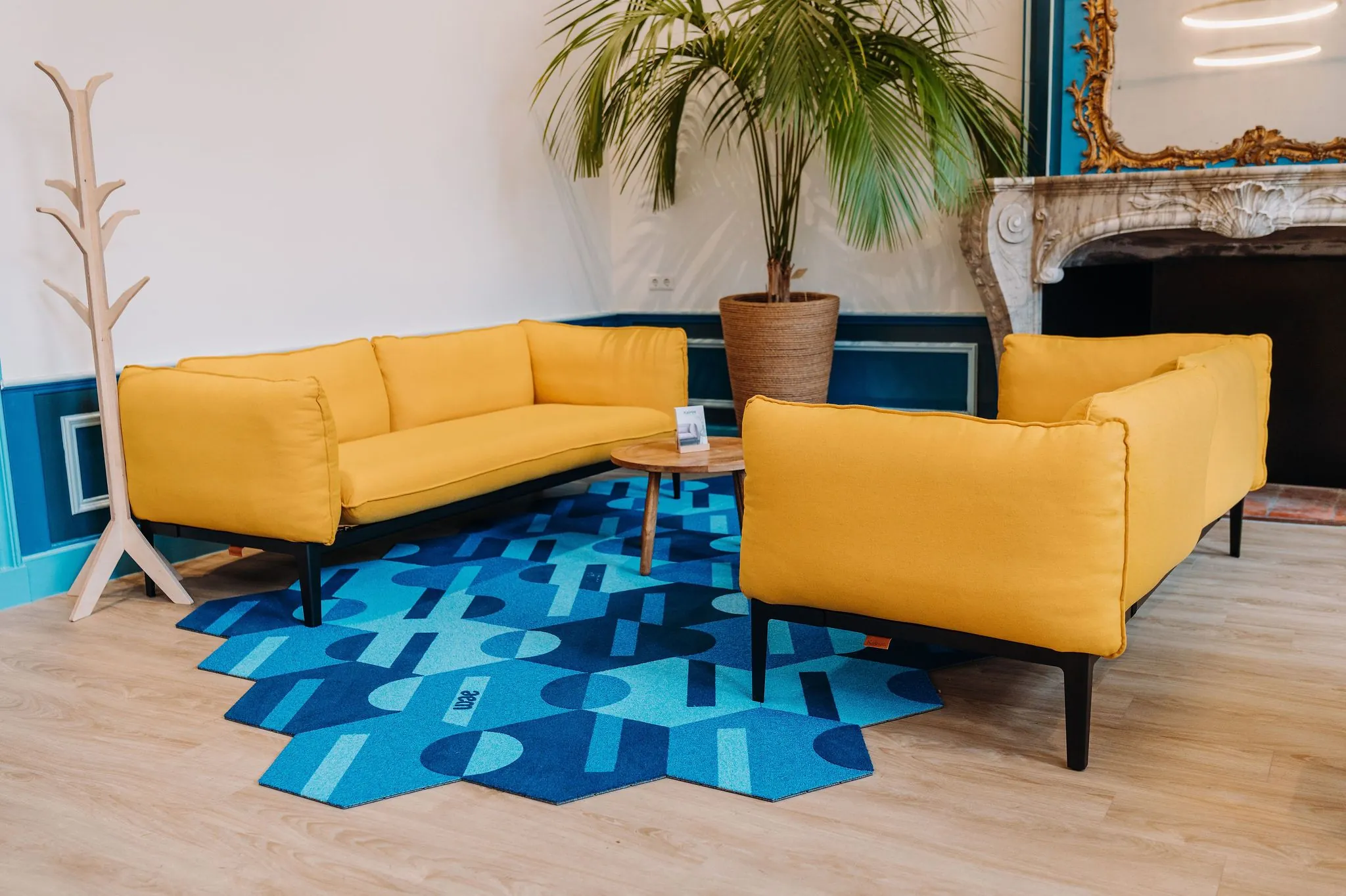 Moderne wachtruimte met twee gele banken, een blauwe vloerkleed, een ronde houten tafel en een grote kamerplant in een kantoor aan de Kromme Nieuwe Gracht in Utrecht.