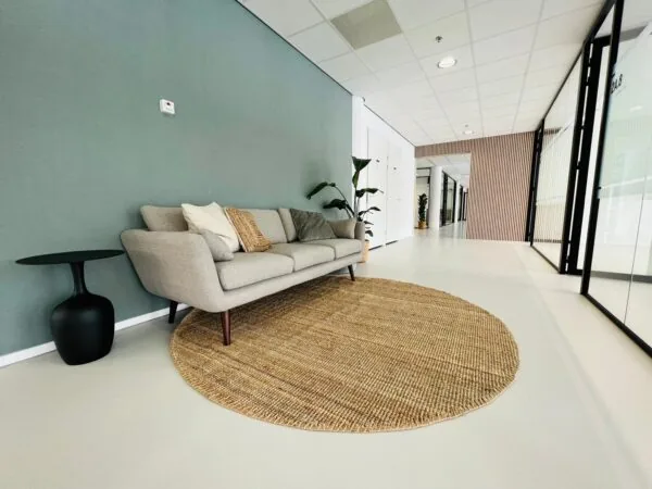 Stijlvolle wachtruimte in een modern kantoor aan het Kruisplein 480 in Rotterdam, met een grijze bank, rond juten vloerkleed en glazen kantoorwanden.