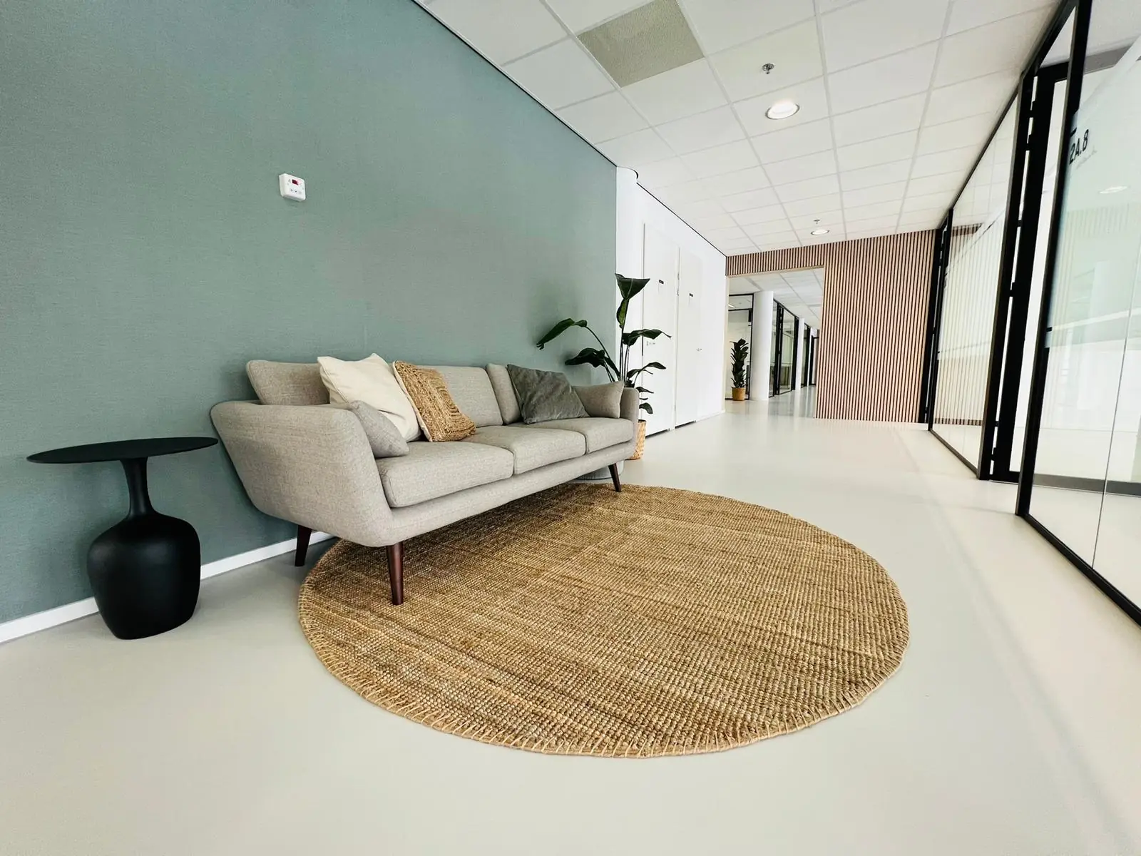 Stijlvolle wachtruimte in een modern kantoor aan het Kruisplein 480 in Rotterdam, met een grijze bank, rond juten vloerkleed en glazen kantoorwanden.