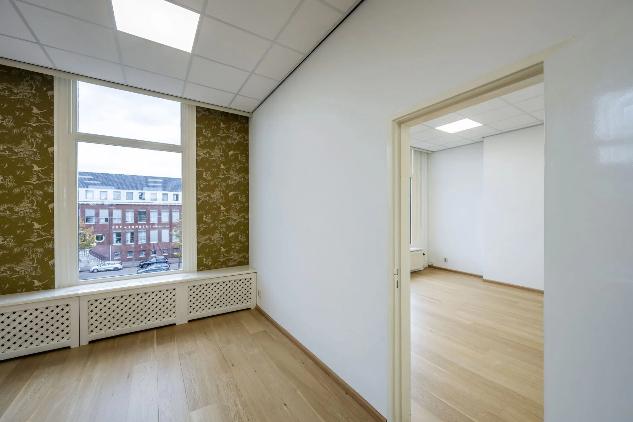 Lichte kantoorruimte aan de Nieuwe Gracht 45-47 in Haarlem met houten vloer, groot raam en doorloop naar aangrenzende kamer.