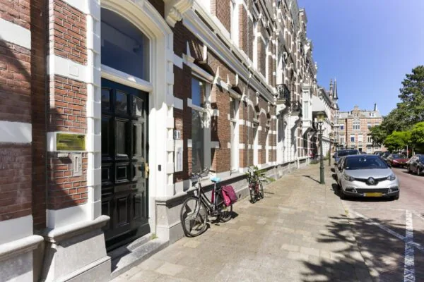 Voorgevel van een klassiek kantoorpand aan het Sweelinckplein in Den Haag met geparkeerde fietsen en auto's.