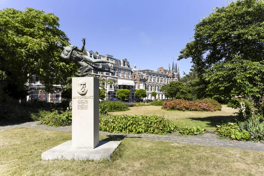 Beeld van een standbeeld van Jan Pieterszoon Sweelinck op een groen plein aan het Sweelinckplein in Den Haag, omringd door historische panden.