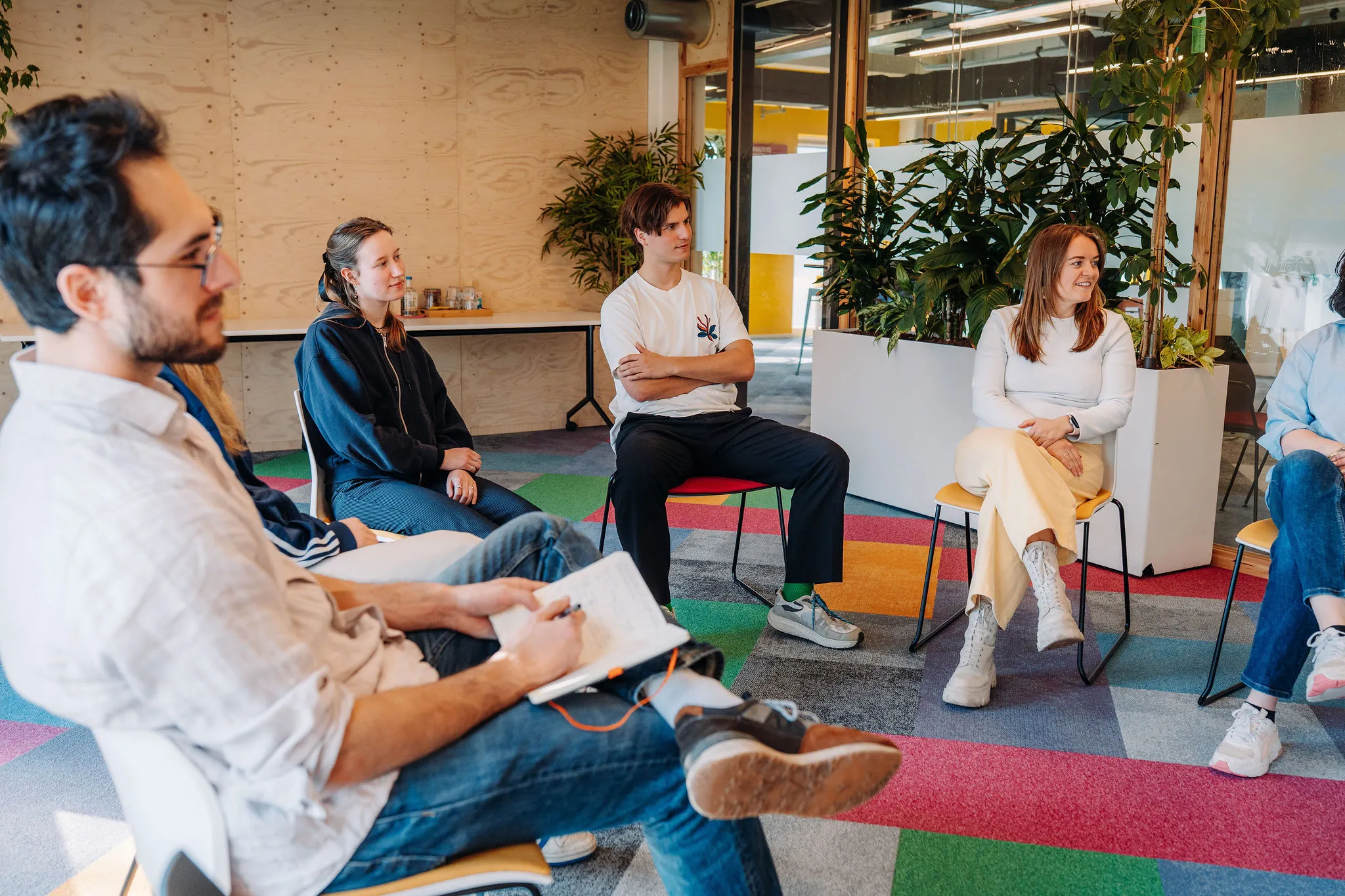 Een groep mensen zit in een kleurrijke kantoorruimte aan de Willem Fenengastraat 14 in Amsterdam in een informele kringbijeenkomst.