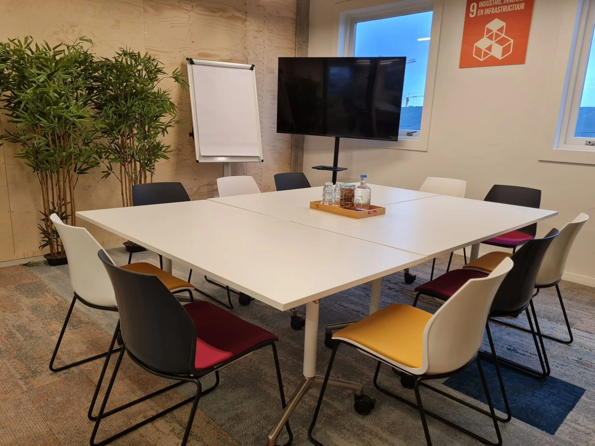 Vergaderruimte aan de Willem Fenengastraat 2 in Amsterdam met een witte tafel, kleurrijke stoelen, een tv-scherm, flip-over en planten.