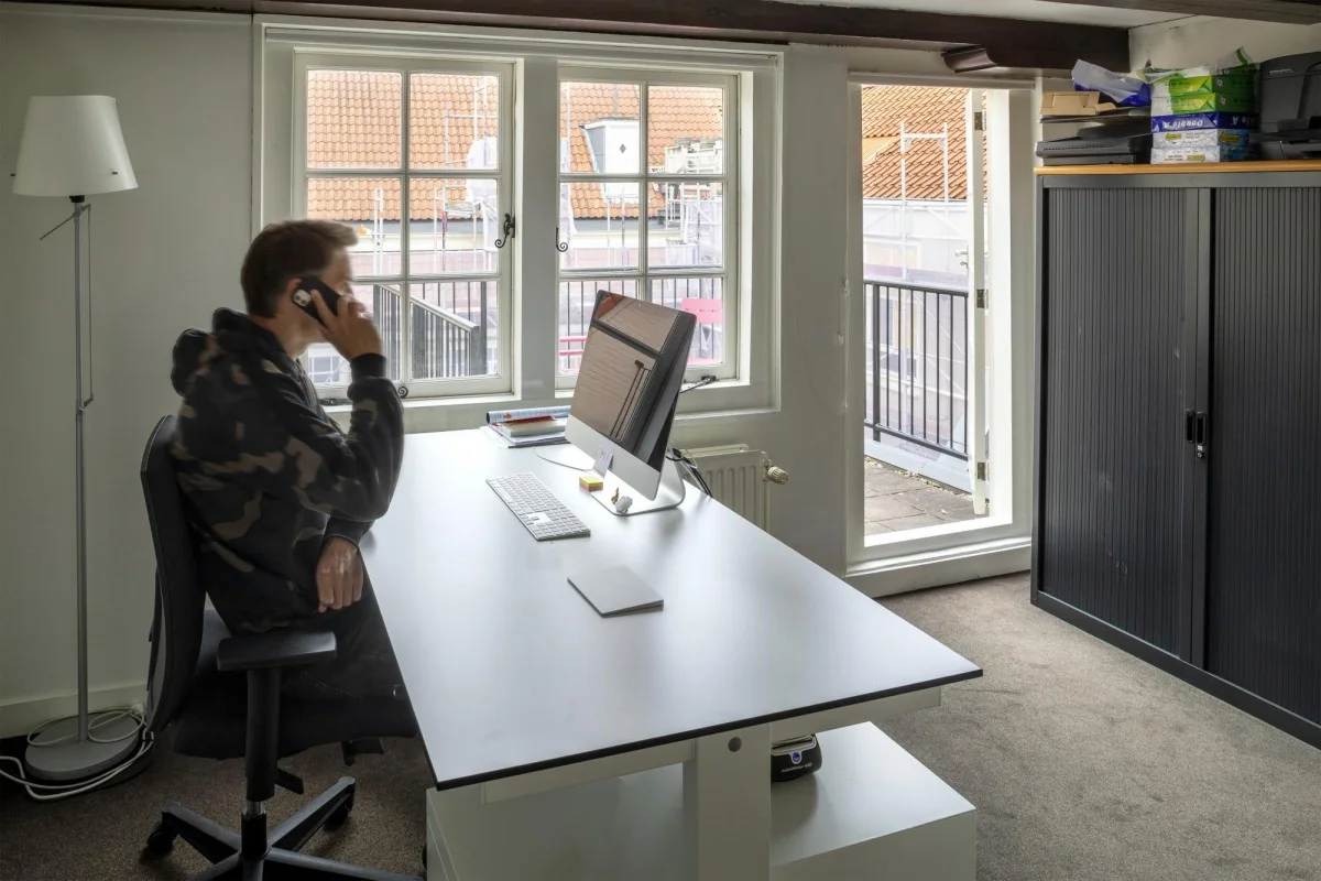 Man aan het werk in een modern kantoor op Keizersgracht 106, zittend aan een bureau en telefonerend bij een raam met uitzicht op een dakterras.