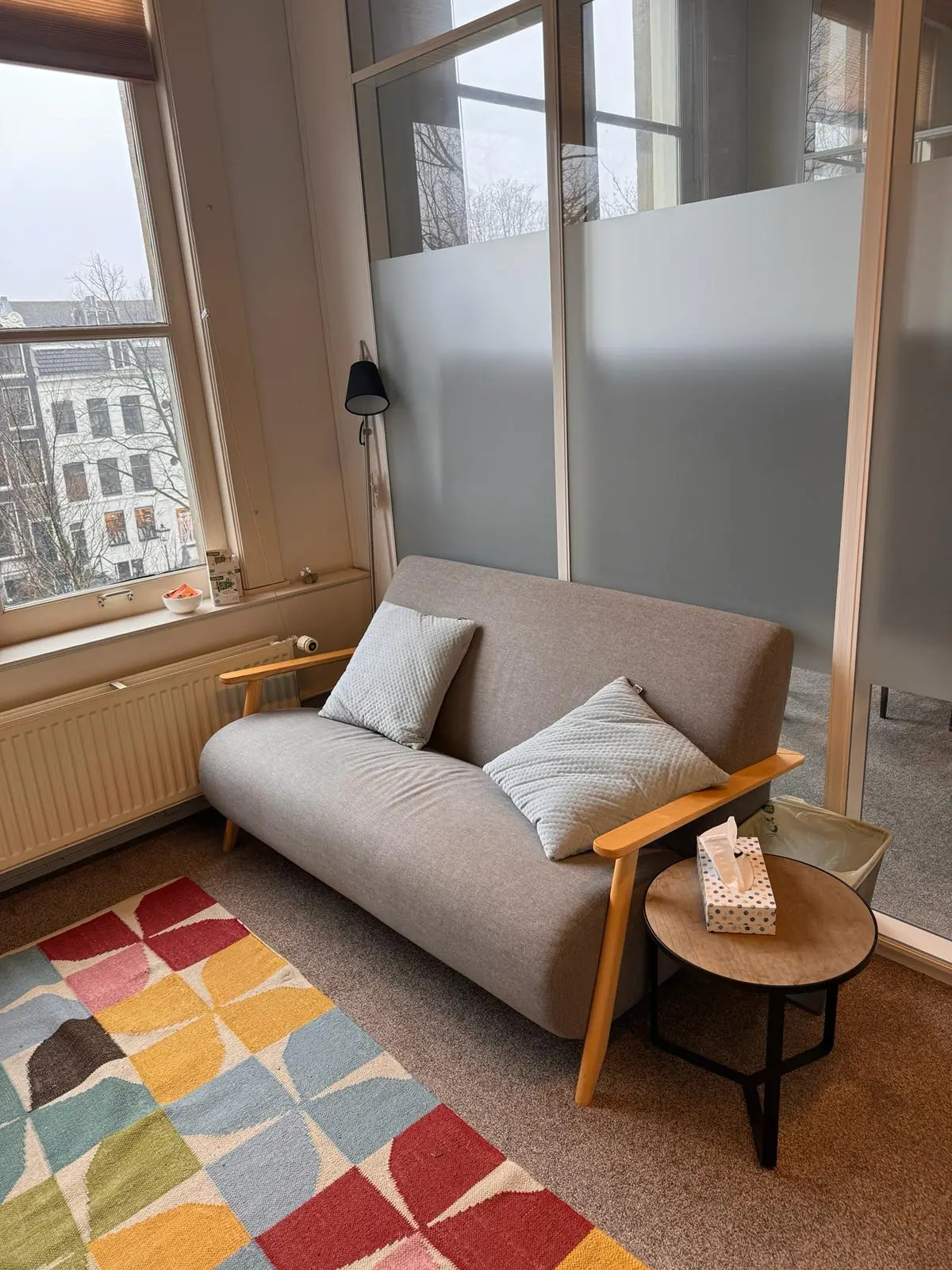 Grijze tweezitsbank met twee kussens naast een tafeltje met tissues in een knusse ruimte aan de Keizersgracht 241.