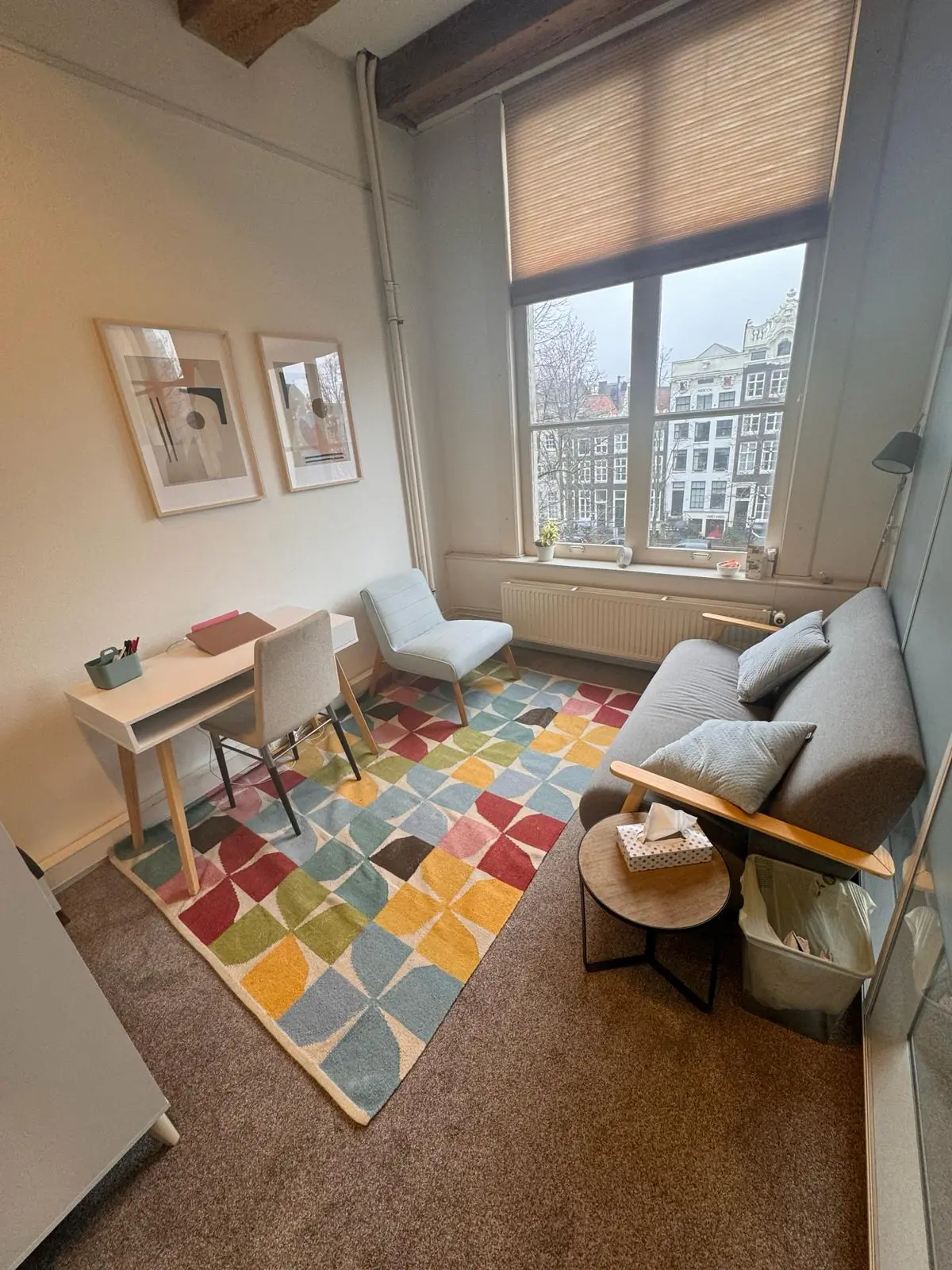 Modern ingerichte kamer aan de Keizersgracht met kleurrijk vloerkleed, bureau, zitbank en uitzicht op Amsterdamse grachtenpanden.
