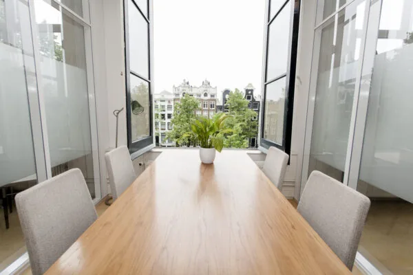 Vergaderruimte aan de Keizersgracht met houten tafel, vier stoelen, open ramen en uitzicht op Amsterdamse grachtenpanden.