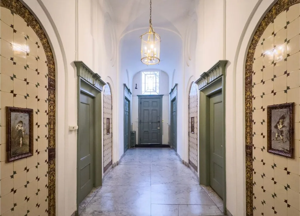 Gedeeltelijk betegelde gang in Keizersgracht 261 met groene deuren, klassieke schilderijen en een lantaarnvormige plafondlamp.