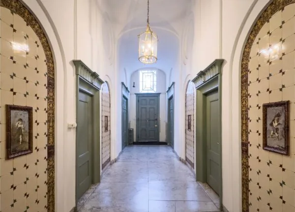 Gedeeltelijk betegelde gang in Keizersgracht 261 met groene deuren, klassieke schilderijen en een lantaarnvormige plafondlamp.