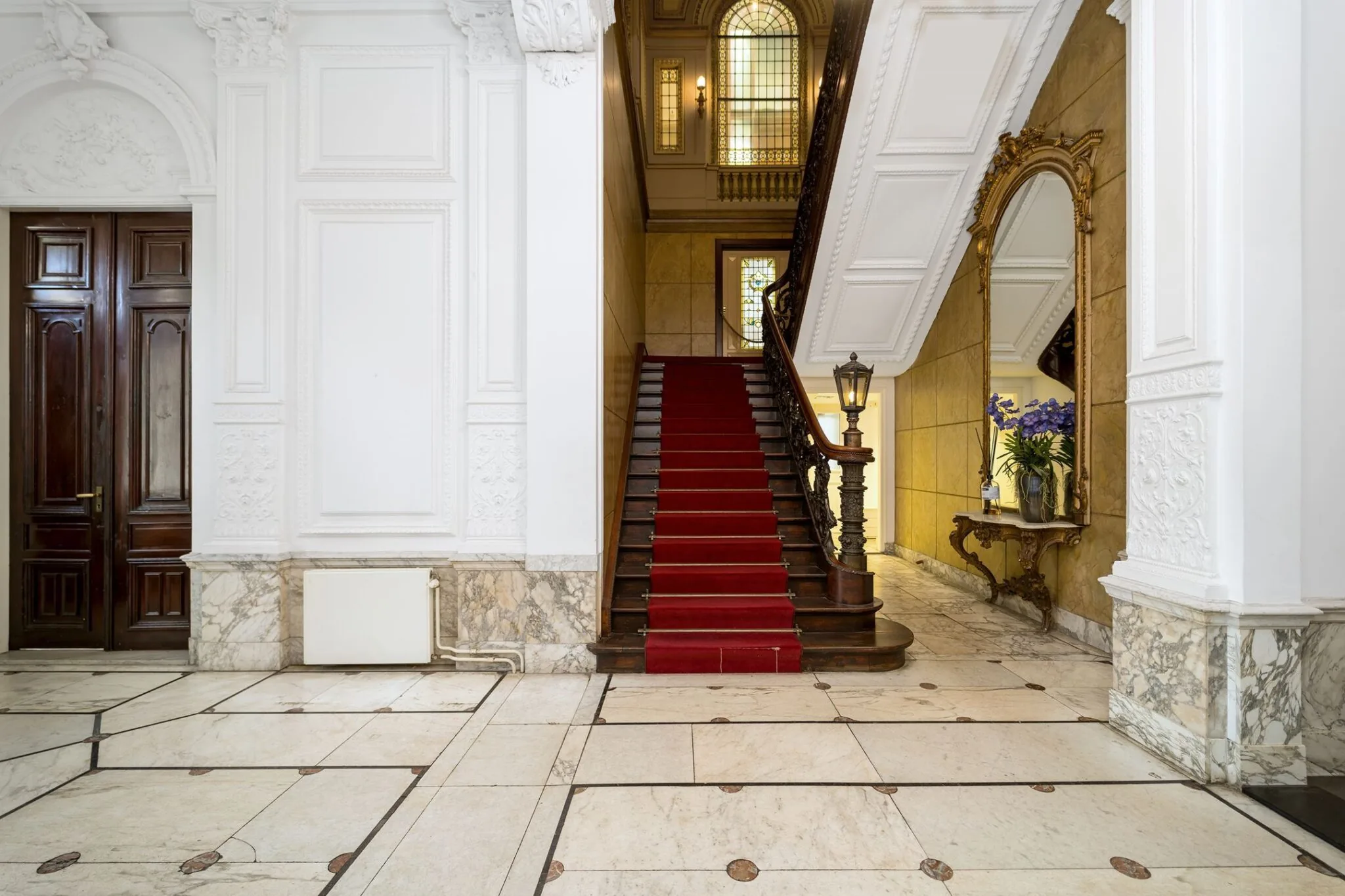 Monumentale hal van Keizersgracht 452 met een houten trap bekleed met rood tapijt, sierlijke details en een grote spiegel.