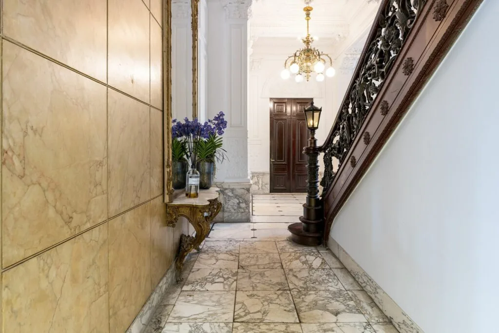 Luxe marmeren hal van Keizersgracht 452 met sierlijke houten trap, kroonluchter en bloemen op een vergulde console.